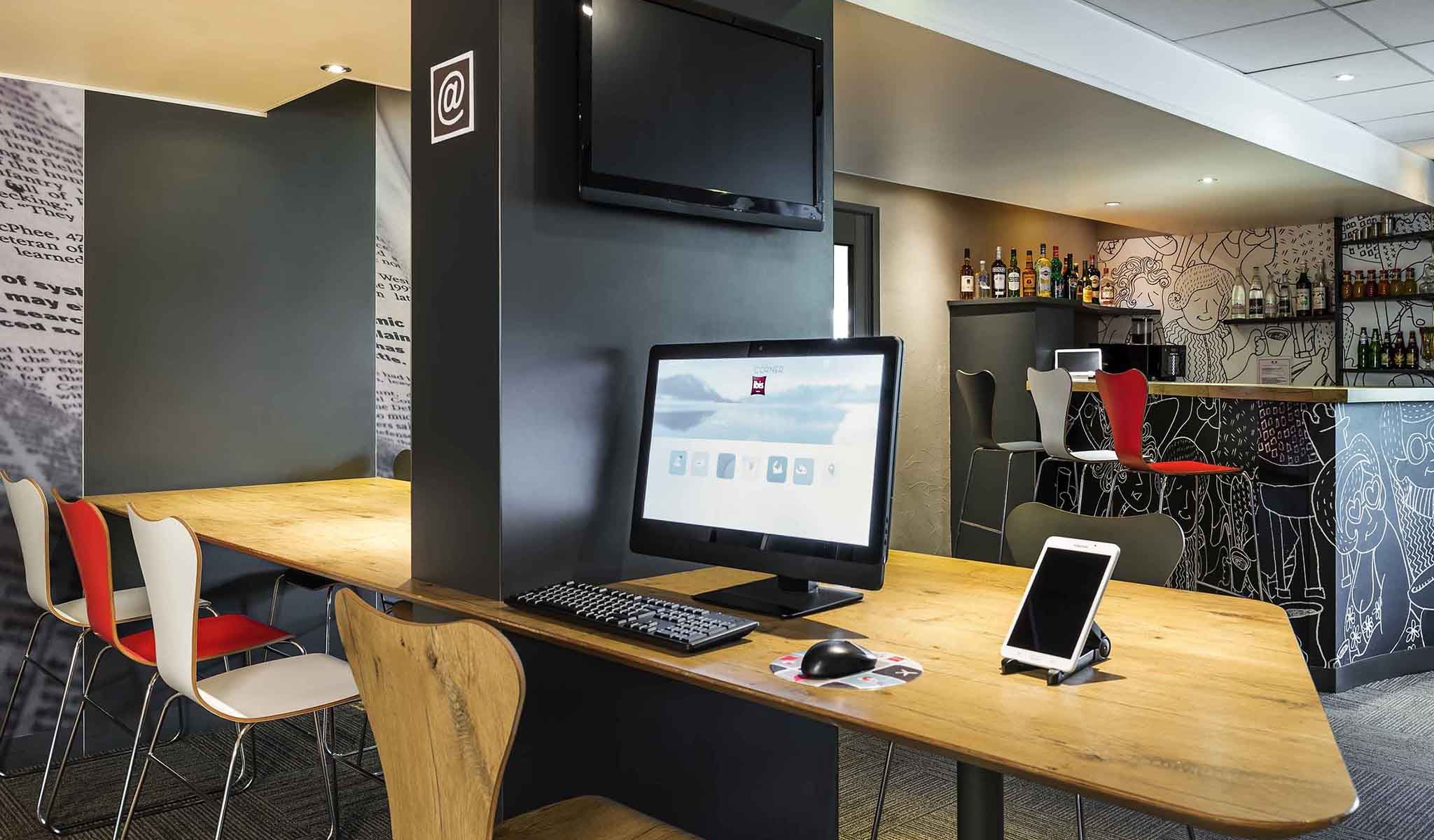 Coin salon d'hôtel idéal pour un séminaire en ville avec une table haute en bois et un poste informatique comprenant un écran noir, un clavier, une souris et une tablette blanche sur support au premier plan, une télévision murale au-dessus et des chaises bistrot rouges et blanches le long du comptoir bar en arrière-plan, sur une moquette grise à motifs.