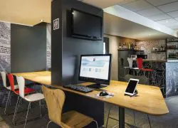 Coin salon d'hôtel idéal pour un séminaire en ville avec une table haute en bois et un poste informatique comprenant un écran noir, un clavier, une souris et une tablette blanche sur support au premier plan, une télévision murale au-dessus et des chaises bistrot rouges et blanches le long du comptoir bar en arrière-plan, sur une moquette grise à motifs.