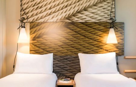 Chambre d'hôtel à lits jumeaux adaptée pour un hébergement groupe avec deux lits simples habillés de linge blanc et d'oreillers au premier plan, une petite table de chevet cubique en bois avec des livres entre les deux lits, deux suspensions luminaires à abat-jour conique suspendues par des câbles noirs de chaque côté, et un grand panneau de tête de lit à motif de corde tressée recouvrant le mur en arrière-plan.
