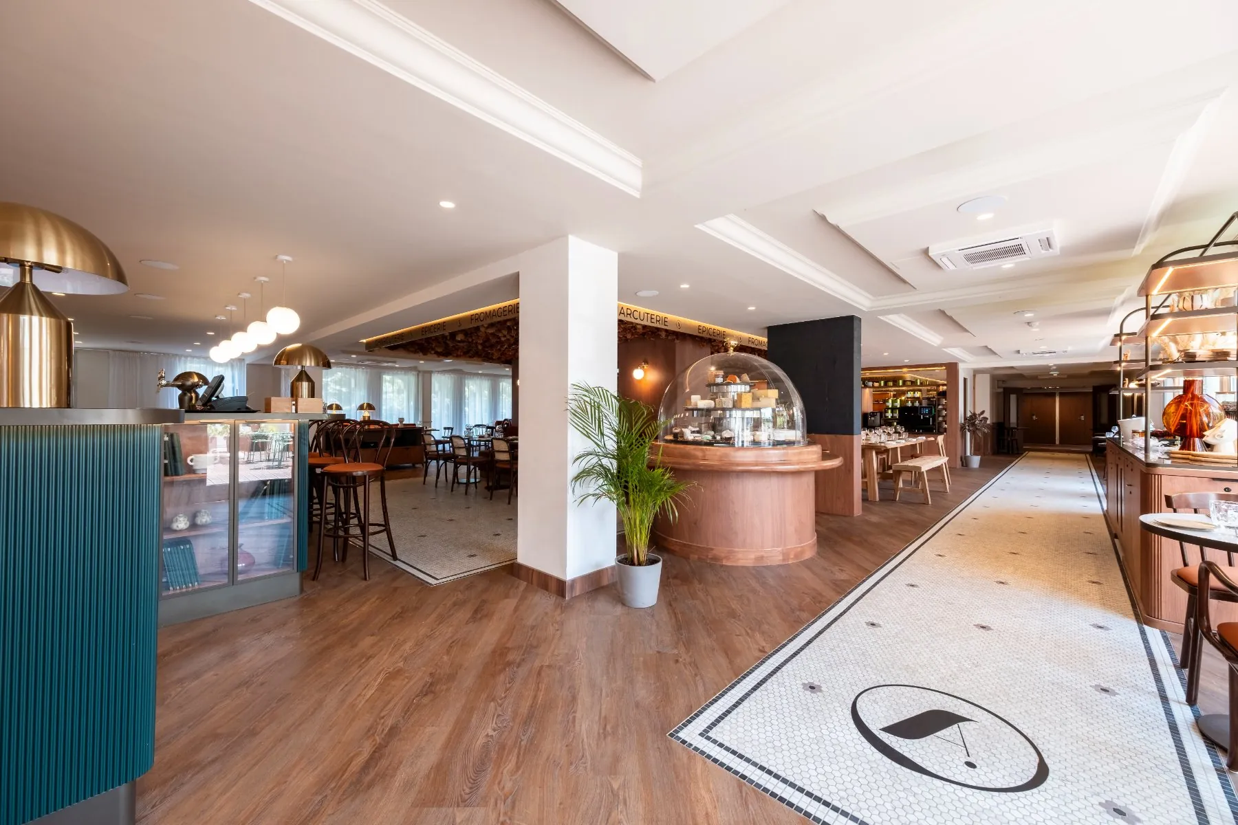 restaurant dining room entrance with, on the right, a bar counter and chairs lined up in front, opposite a giant cheese bell, a communal table, set tables, and on the right a wooden and brass buffet