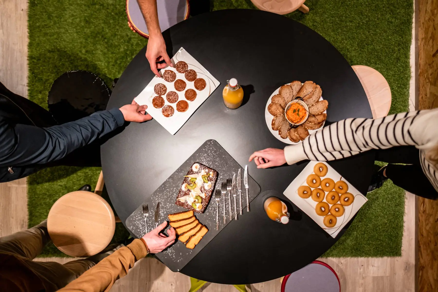 en::Black round table with cakes displayed on slate boards, doughnuts, toasted bread with sauces, fruit juices and small desserts, with guests serving themselves around the table. fr::Table ronde noire avec cakes et gâteaux présentés sur ardoise, beignets, tartines grillées et sauces d’accompagnement, jus de fruits et petits desserts, avec des personnes autour en train de se servir.