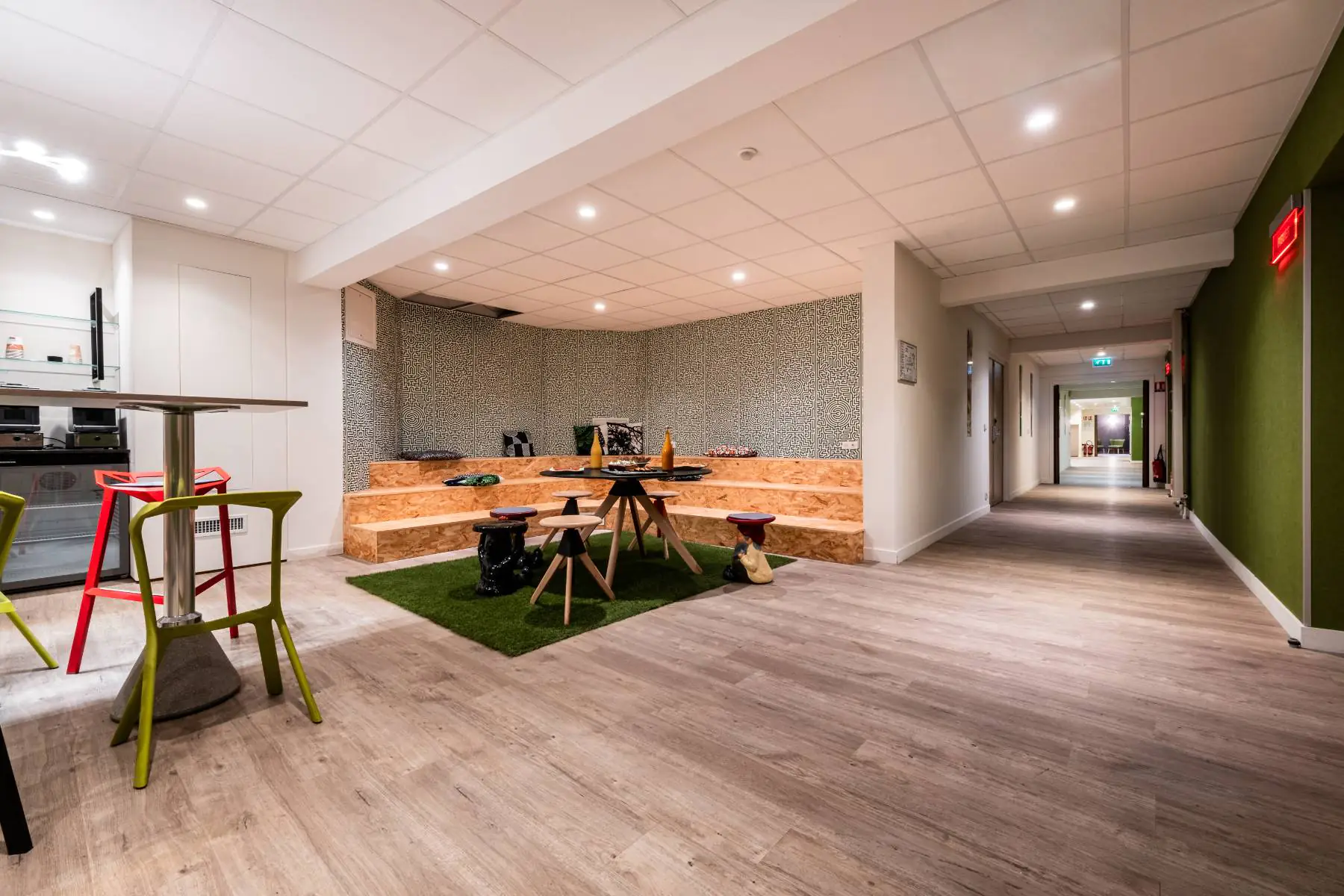 en::Break area with a central high table and colorful bar stools, corner spaces featuring natural-material platforms with design tables and stools, and a view of the corridor leading to the meeting rooms. fr::Espace pause avec table haute centrale et chaises de bar colorées, espaces en angle aménagés avec estrades en matière naturelle, tables et tabourets design, et vue sur le couloir menant aux salles de réunion.