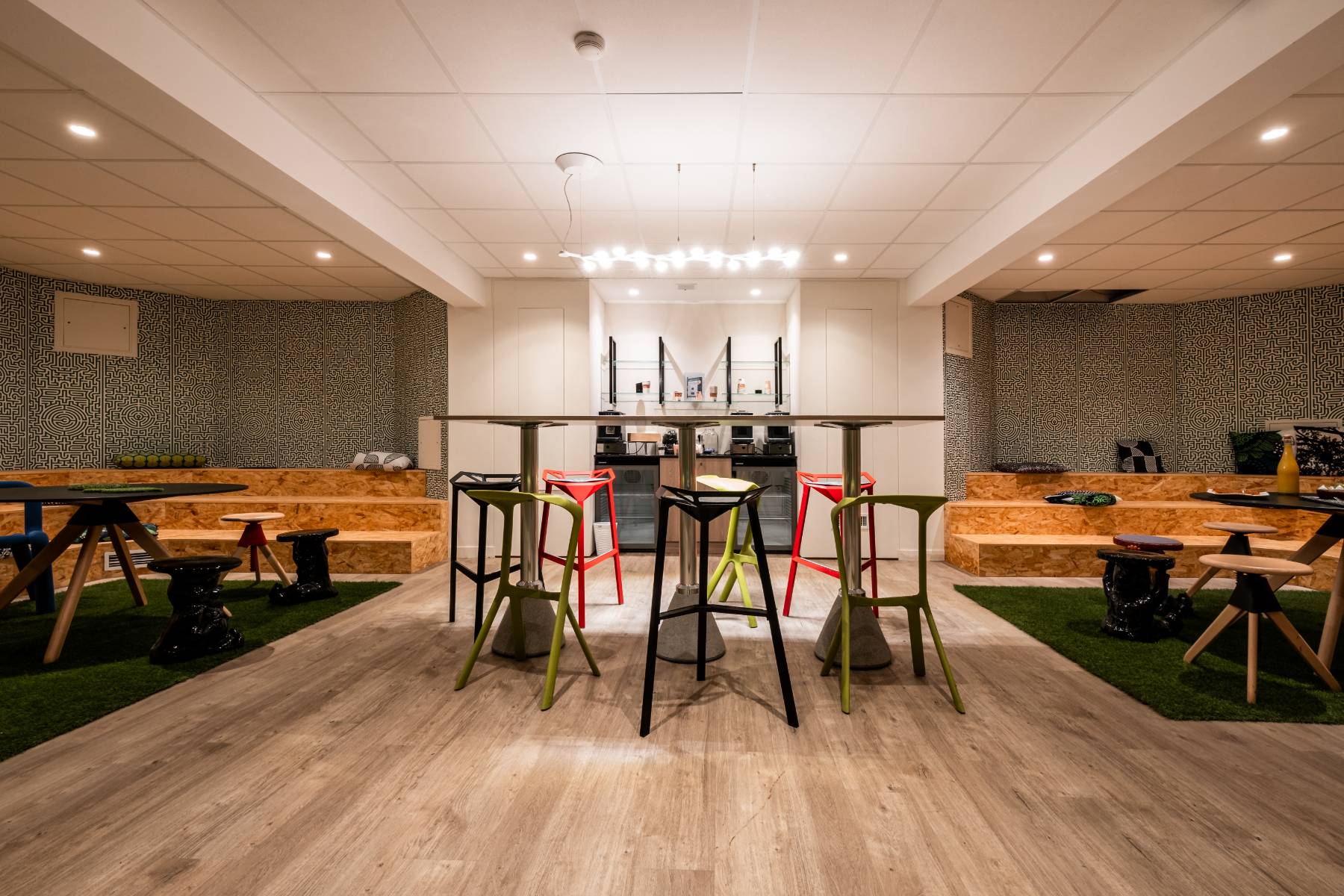 en::Central high table with colorful bar stools, and on each side corner areas featuring natural-material platforms with design tables and stools. fr::Table haute centrale avec chaises de bar colorées, et de chaque côté des espaces en angle aménagés avec estrades en matière naturelle, tables et tabourets design.