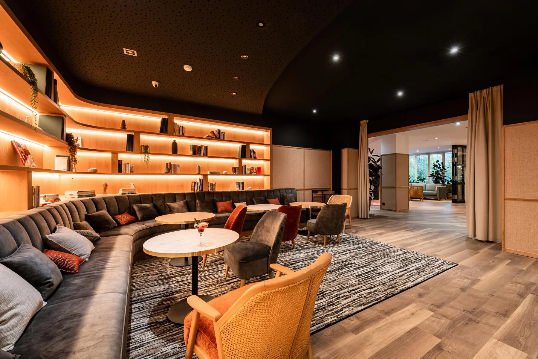 en::library area extending from the lobby and bar, partially visible, with backlit shelves, sofas and lounge seating fr::bibliothèque dans le prolongement du lobby et du bar, partiellement visible, avec étagères rétroéclairées, canapés en tissu et velours, table ronde et fauteuils devant