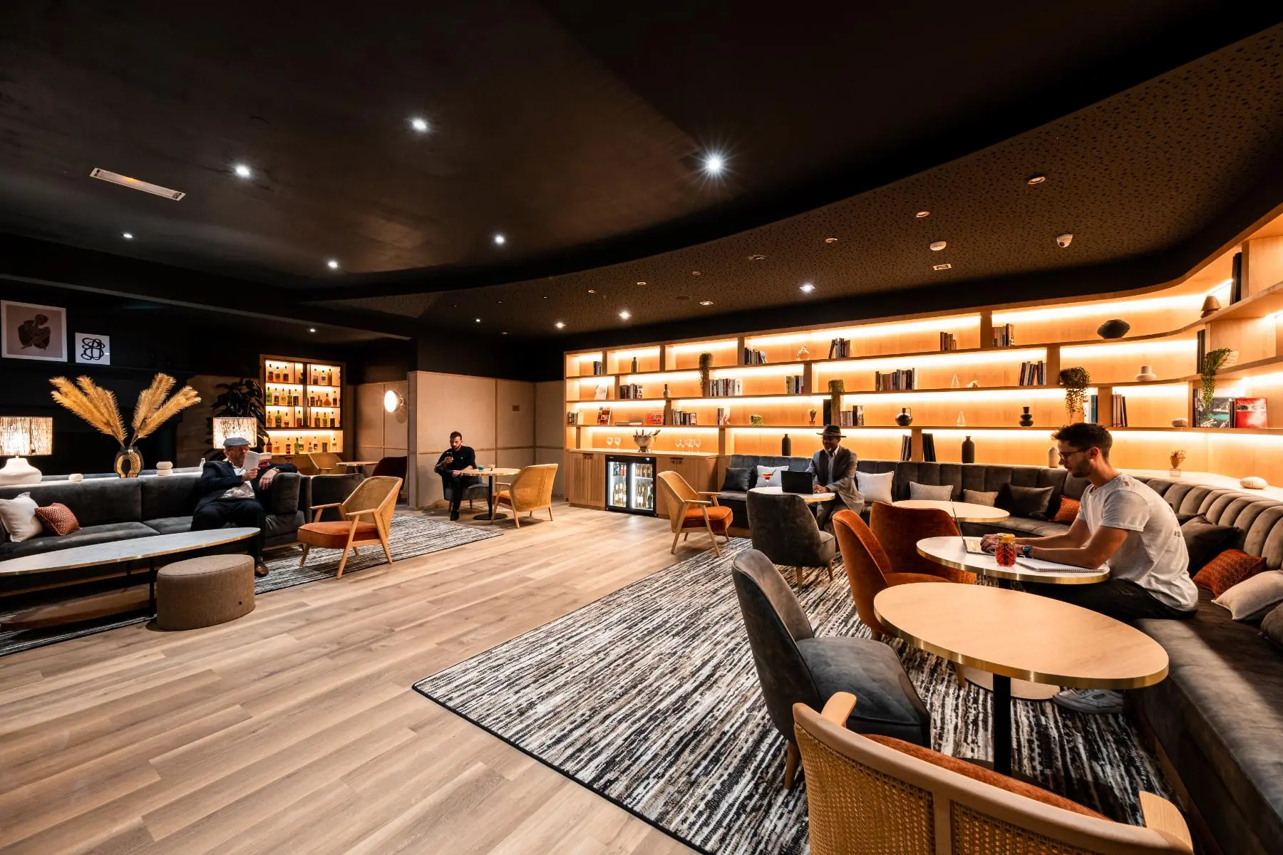 en::library-style work and lounge area next to the bar, with a three-level backlit angular bookshelf, grey sofas, tables and wicker armchairs, guests working, reading or enjoying a drink fr::espace de travail ou de détente à côté du bar, avec bibliothèque angulaire rétroéclairée sur trois niveaux, canapés gris, tables et fauteuils en osier, personnes assises en train de travailler, lire ou boire un verre
