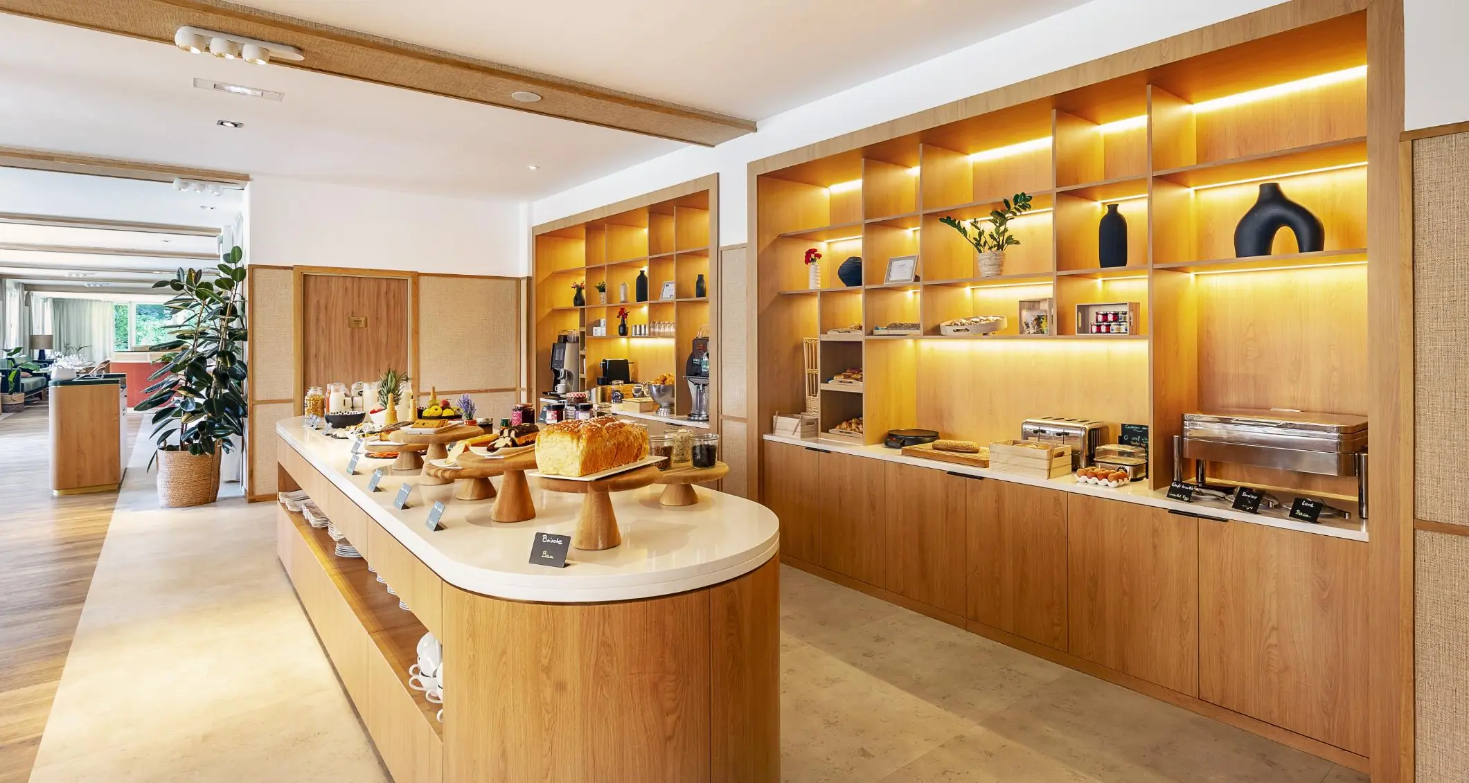 en::breakfast buffet with a rounded wooden design and white finish, backlit wooden shelves behind, offering pastries, savory and sweet items, milk jars and fruit juices fr::buffet petit déjeuner au design arrondi en bois et revêtement blanc, avec étagères en bois rétroéclairées à l’arrière, proposant pâtisseries, mets salés et sucrés, pots de lait et jus de fruits