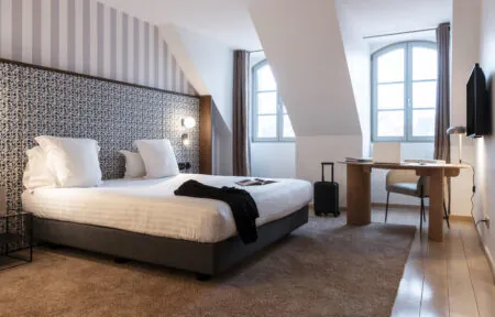 en::Double bed with a white duvet and two rows of white pillows, wooden desk with a modern rounded beige armchair, black jacket on the bed with an open book, wall-mounted television and lights switched on, brown rug over wooden flooring, and a floral fabric bed frame matching the striped wall covering. fr::Lit double avec couette blanche et double rangée d’oreillers blancs, bureau en bois avec fauteuil beige aux lignes arrondies, veste noire posée sur le lit avec livre ouvert, télévision murale et luminaires allumés, tapis marron sur parquet, encadrement de lit en tissu fleuri assorti à la tapisserie murale rayée.