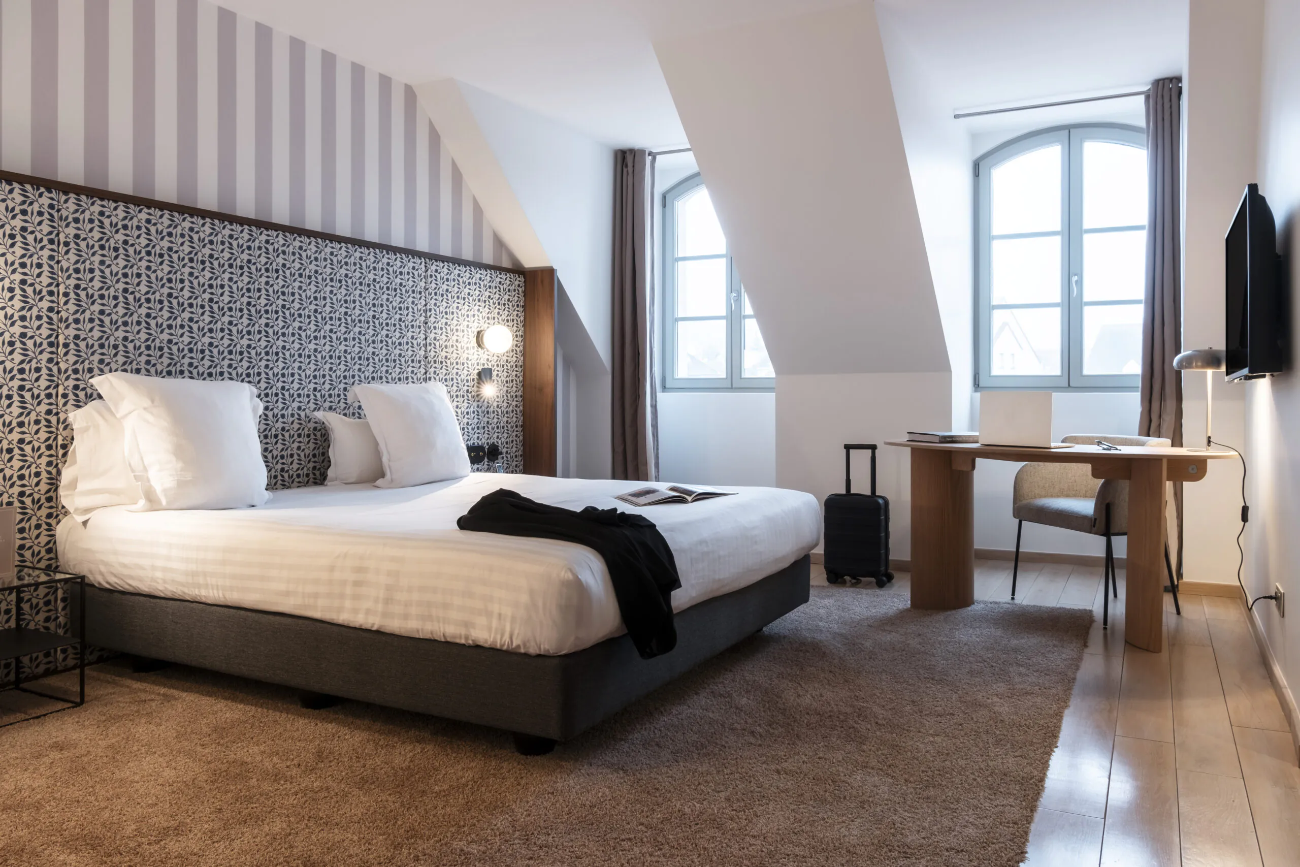 en::Double bed with a white duvet and two rows of white pillows, wooden desk with a modern rounded beige armchair, black jacket on the bed with an open book, wall-mounted television and lights switched on, brown rug over wooden flooring, and a floral fabric bed frame matching the striped wall covering. fr::Lit double avec couette blanche et double rangée d’oreillers blancs, bureau en bois avec fauteuil beige aux lignes arrondies, veste noire posée sur le lit avec livre ouvert, télévision murale et luminaires allumés, tapis marron sur parquet, encadrement de lit en tissu fleuri assorti à la tapisserie murale rayée.