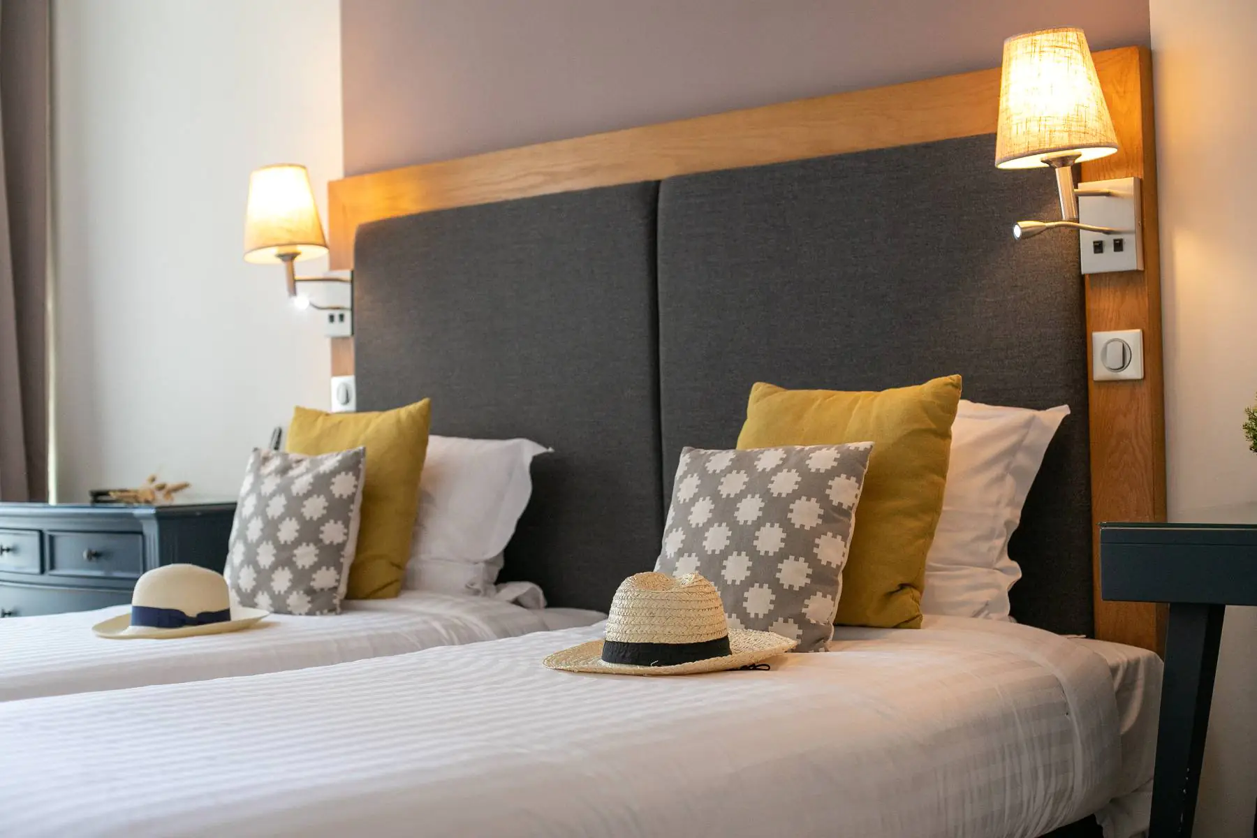en::Room with two twin beds dressed in white duvets, grey, white and mustard cushions, a straw hat placed on each bed, wooden headboard with grey fabric panels, wall lights and reading lamps switched on, and a chest of drawers and bedside tables framing the beds. fr::Chambre avec deux lits jumeaux aux couettes blanches, coussins gris, blancs et moutarde, chapeau de paille posé sur chaque lit, tête de lit en bois avec revêtement en tissu gris, appliques et liseuses allumées, commode et tables de chevet encadrant l’ensemble.