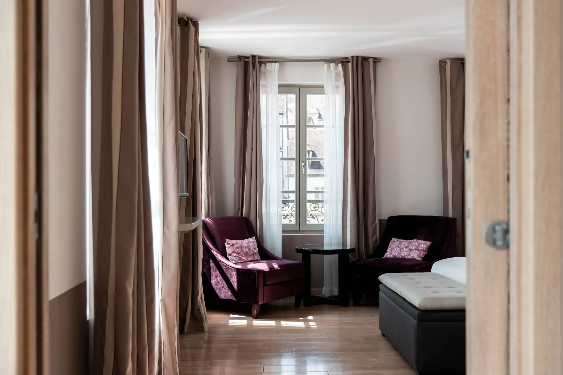 en::View of a bedroom from the doorway, showing the foot of the bed with a cushioned bench, mauve velvet armchairs with matching cushions, and windows framed by white sheer curtains and beige double drapes letting light in over the wooden floor. fr::Vue d’une chambre depuis la porte, avec le pied du lit et un coffre d’assise garni d’un coussin, fauteuils en velours mauve assortis, fenêtres encadrées de voilages blancs et de doubles rideaux beiges laissant entrer la lumière sur le parquet.