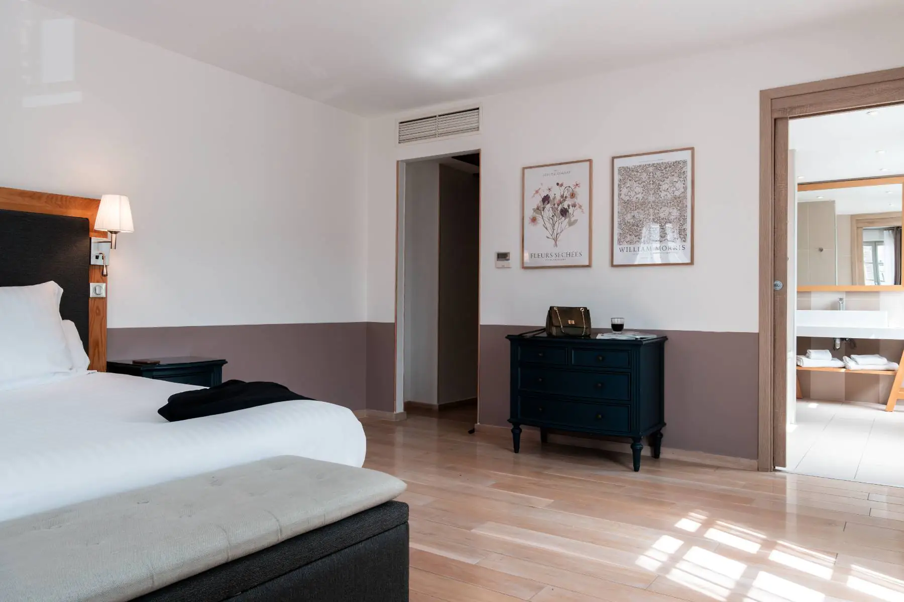 en::Room with a double bed, antique-style chest of drawers, lithographs on the wall, and an open doorway leading to the bathroom with a wide sink and full-length mi fr::Chambre avec lit double, commode d’inspiration ancienne, lithographies au mur, porte d’accès ouverte sur la salle de bain avec large lavabo et miroir pleine longueur.