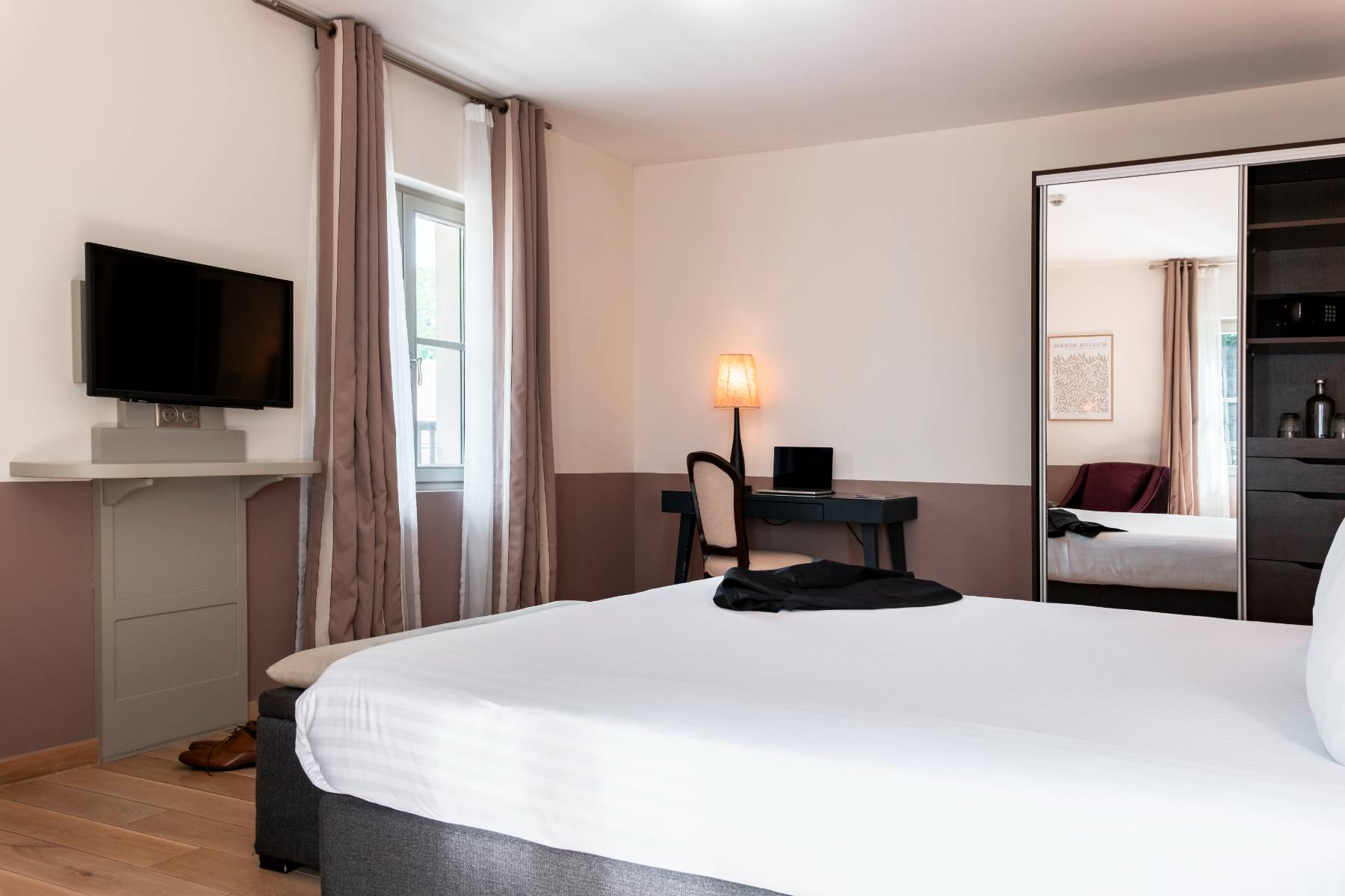 en::Room with a double bed, wall-mounted television, desk with a computer and a lit lamp, antique chair, open wardrobe with a visible safe, and a suit jacket placed on the bed. fr::Chambre avec lit double, télévision murale, bureau équipé d’un ordinateur et d’une lampe allumée, chaise ancienne, armoire ouverte avec coffre-fort visible, et veste de costume posée sur le lit.
