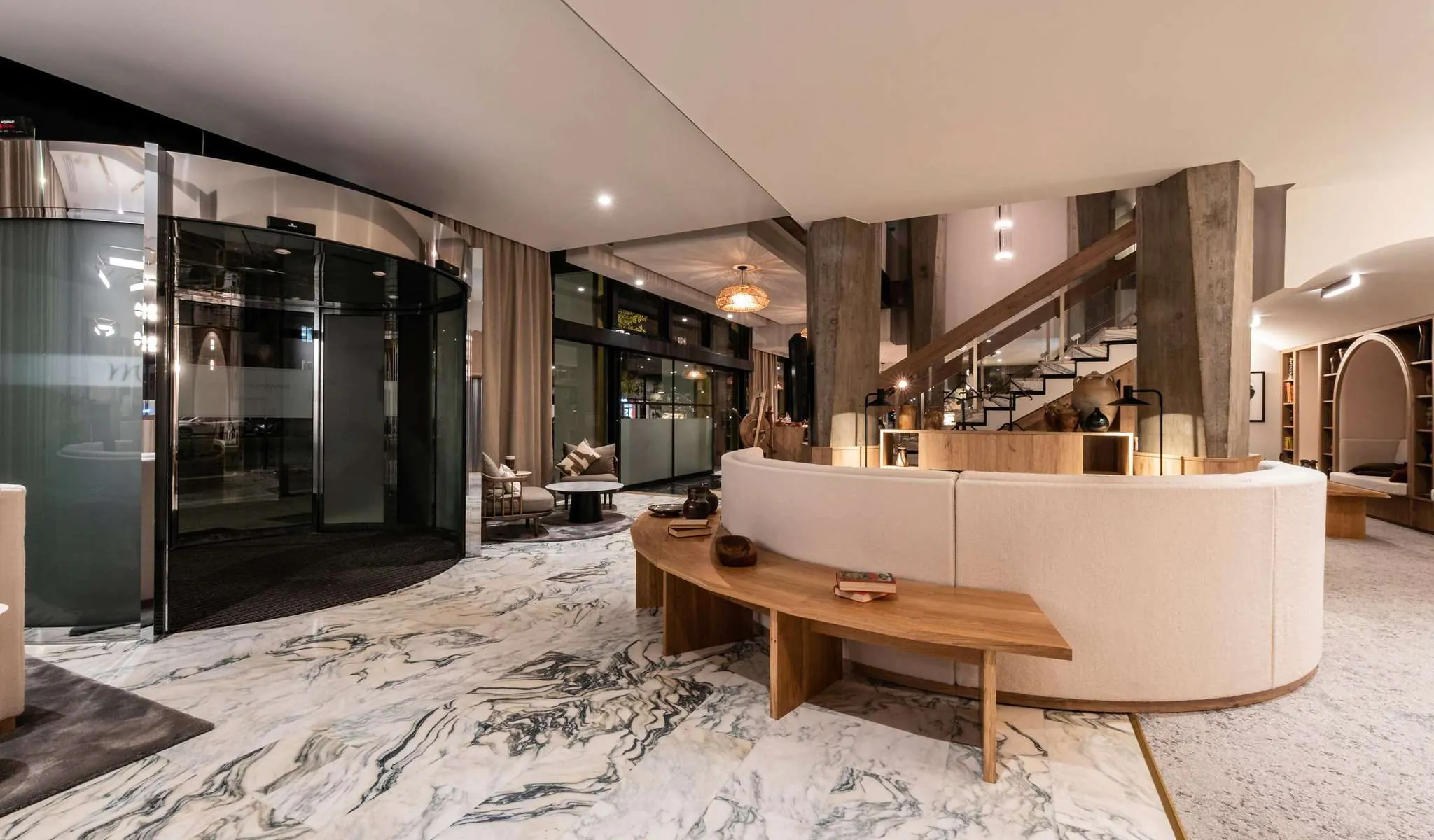 Hall d'hôtel adapté pour un séminaire en ville, comprenant un sol en marbre veiné blanc et gris au premier plan, un canapé arrondi crème avec banc en bois au centre, une porte tambour noire à gauche, et un escalier en béton avec rambarde en bois menant à l'étage en arrière-plan.