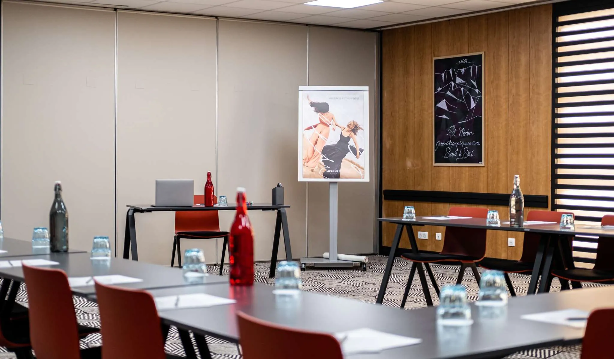 Salle de formation installée en format U, comprenant des tables noires avec chaises orange et bouteilles d'eau disposées au premier plan, un ordinateur portable sur une table d'appoint au centre, une affiche sur pied, et une œuvre encadrée à thème montagne sur un mur en panneaux de bois en arrière-plan.