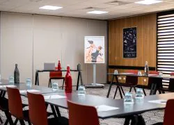 Training room set up in a U-shaped layout, featuring black tables with orange chairs and water bottles arranged in the foreground, a laptop on a side table in the centre, a poster on a stand, and a mountain-themed framed artwork on a wooden panelled wall in the background.