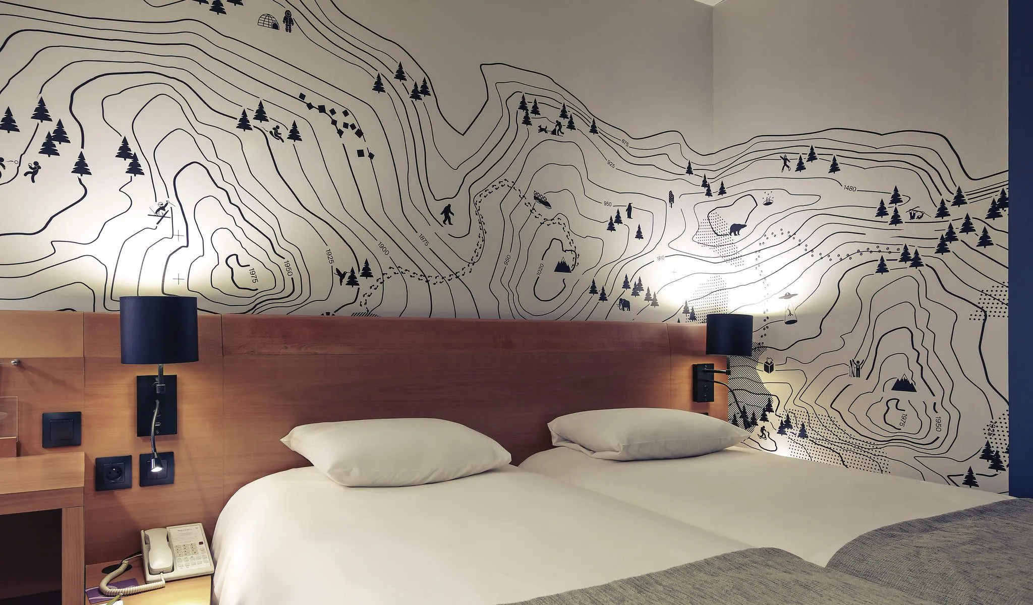 Chambre d'hôtel évoquant un séminaire à la montagne, comprenant un lit blanc avec plaid gris et deux oreillers au premier plan, une tête de lit en bois avec lampes noires et liseuses, un téléphone sur la table de chevet à gauche, et un mur recouvert d'une illustration de courbes de niveau montagneuses en noir et blanc avec skieurs, arbres et animaux en arrière-plan.