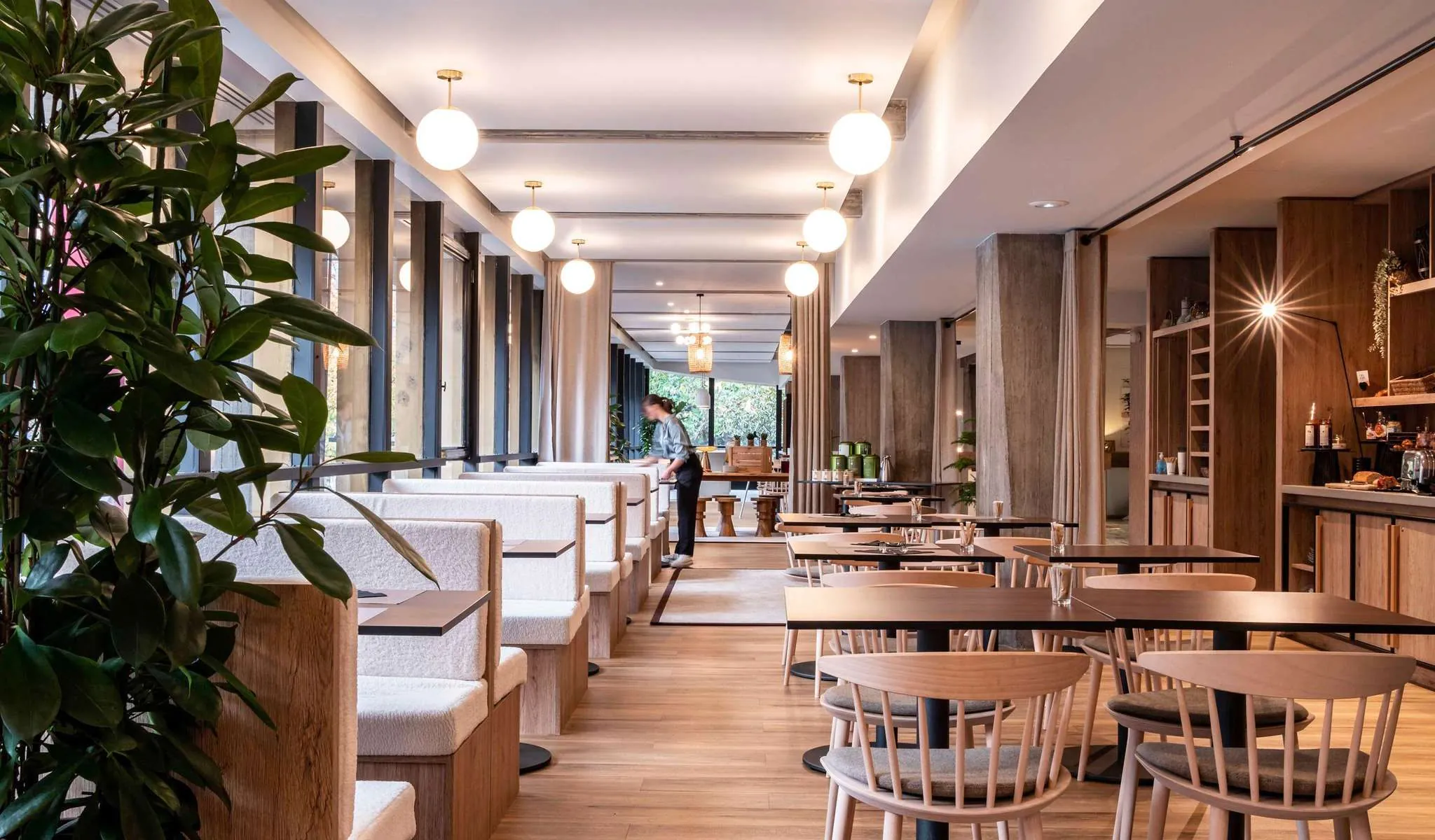 Restaurant d'hôtel proposant une restauration sur place, comprenant une grande plante verte au premier plan à gauche, des tables en bois avec chaises claires à barreaux et petites tables sombres avec banquettes blanches sur un sol en bois au centre, un membre du personnel debout près d'un comptoir bar, et des suspensions lumineuses en verre rond au plafond.