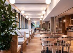 Hotel restaurant offering on-site catering, featuring a large green plant in the foreground on the left, wooden tables with light spindle chairs and small dark tables with white banquette seating on a wooden floor in the centre, a staff member standing near a bar counter, and round glass pendant lights hanging from the ceiling.