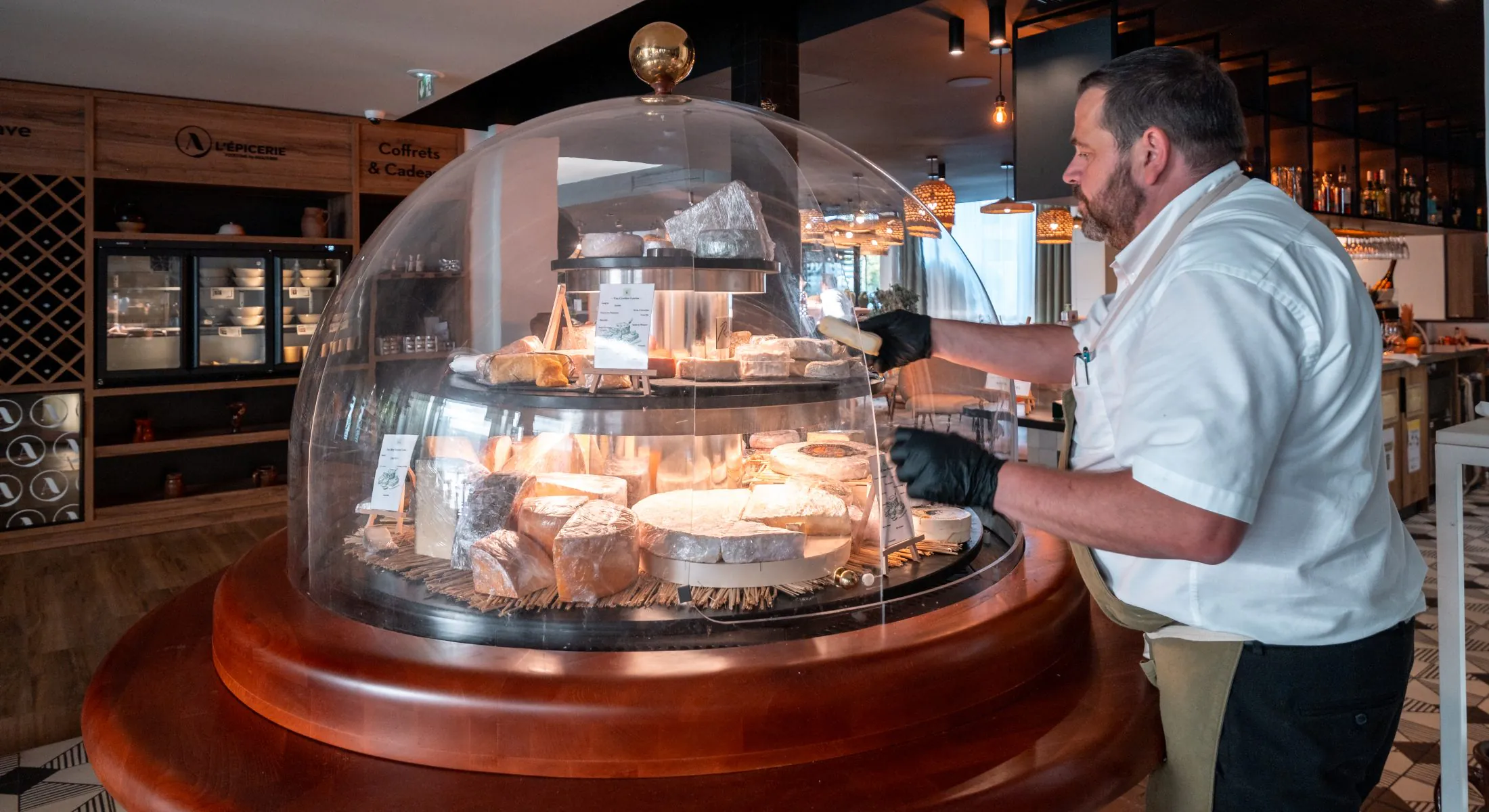 en::1.80 m cheese dome in front of the À l’Épicerie Restaurant Experience gourmet shop, with a person serving cheese. fr::Cloche à fromage de 1,80 m devant la boutique épicerie « À l’Épicerie Restaurant Experience », avec une personne servant du fromage.
