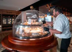 en::1.80 m cheese dome in front of the À l’Épicerie Restaurant Experience gourmet shop, with a person serving cheese. fr::Cloche à fromage de 1,80 m devant la boutique épicerie « À l’Épicerie Restaurant Experience », avec une personne servant du fromage.