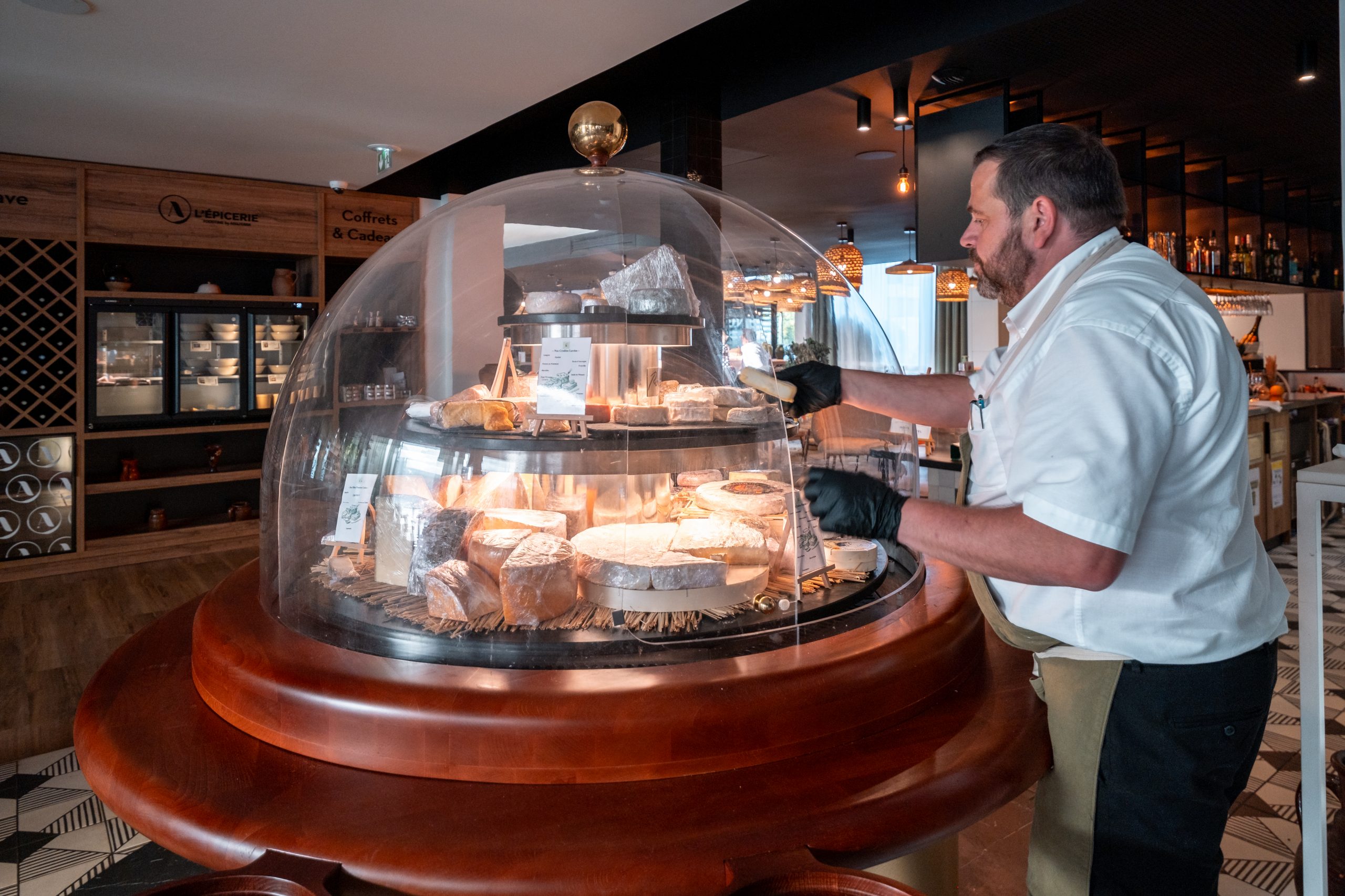 en::1.80 m cheese dome in front of the À l’Épicerie Restaurant Experience gourmet shop, with a person serving cheese. fr::Cloche à fromage de 1,80 m devant la boutique épicerie « À l’Épicerie Restaurant Experience », avec une personne servant du fromage.
