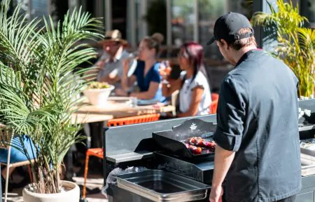 en::Chef in a black uniform and cap, seen from behind, preparing a barbecue in front of guests eating and drinking in the shade of greenery, facing the seminar restaurant’s bay windows. fr::Cuisinier en tenue noire et casquette, vu de dos, préparant un barbecue devant des clients qui mangent et boivent à l’ombre de la végétation, face aux baies vitrées du restaurant séminaire.
