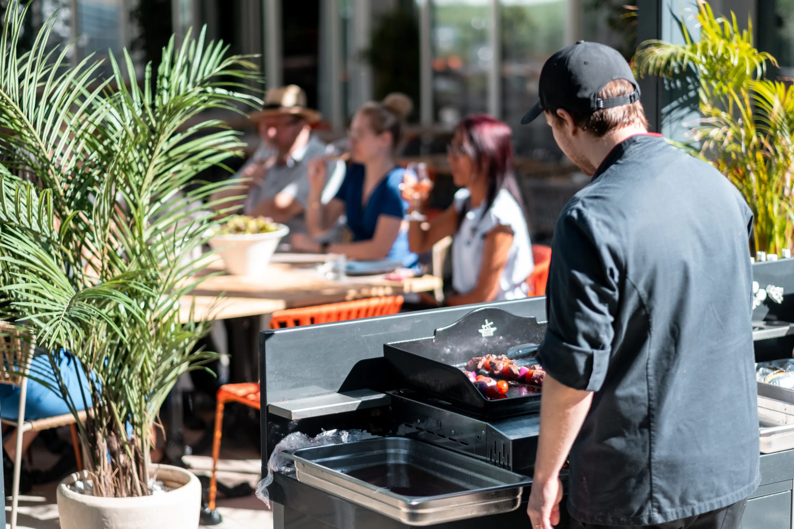 en::Chef in a black uniform and cap, seen from behind, preparing a barbecue in front of guests eating and drinking in the shade of greenery, facing the seminar restaurant’s bay windows. fr::Cuisinier en tenue noire et casquette, vu de dos, préparant un barbecue devant des clients qui mangent et boivent à l’ombre de la végétation, face aux baies vitrées du restaurant séminaire.