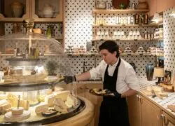 a man in an apron holds a tray of cheese in front of a glass dome