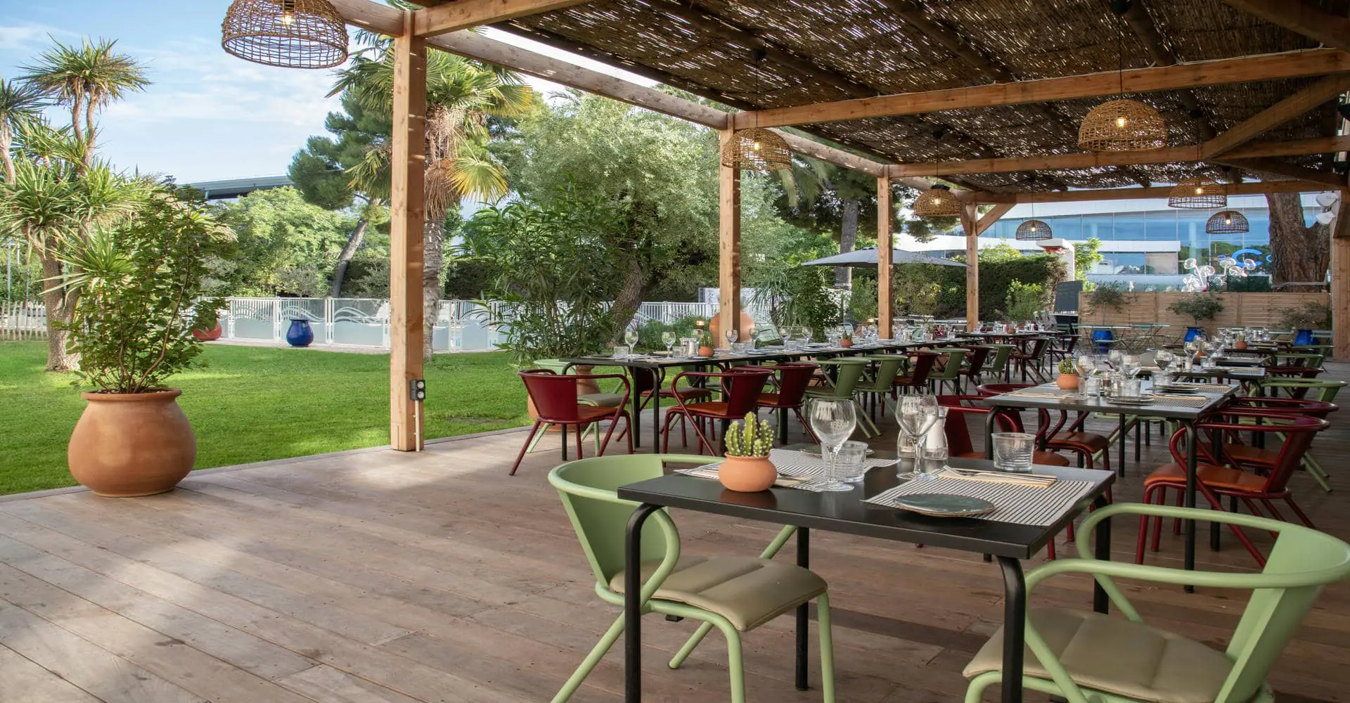 Une terrasse de restaurant en extérieur avec des tables dressées, qui donne sur une pelouse et une piscine à l'arrière plan.