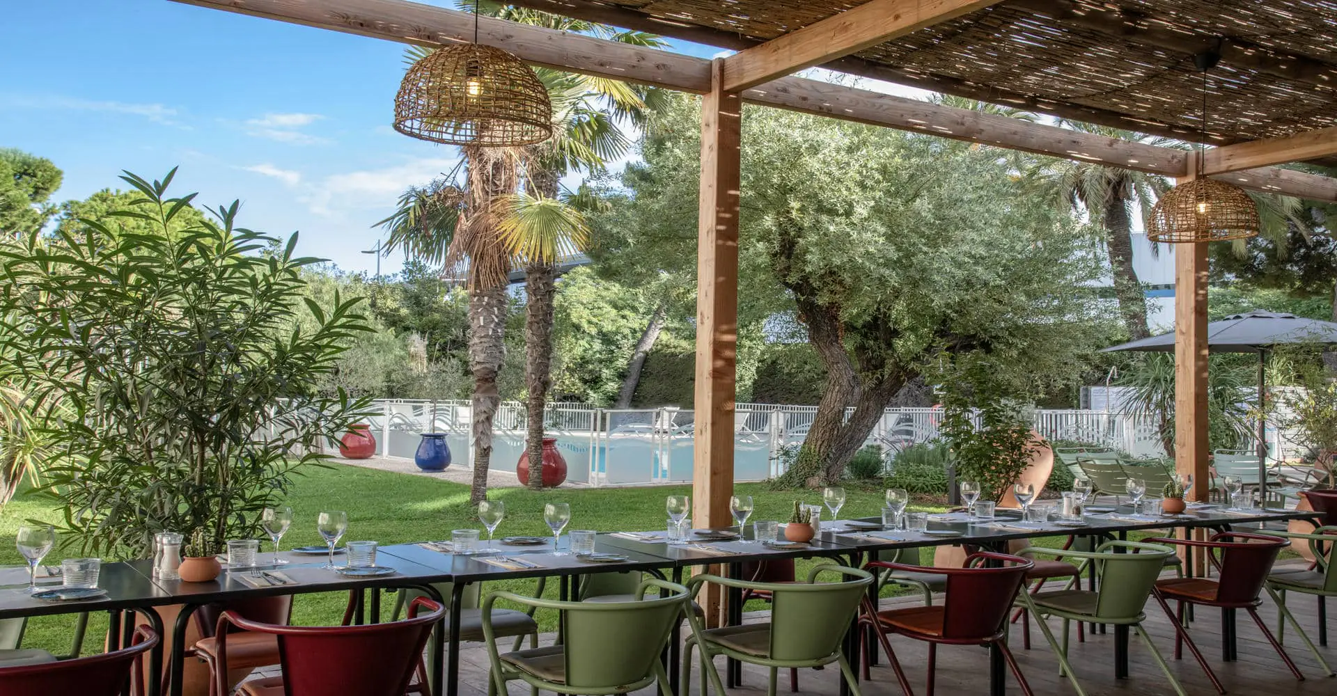 Une terrasse de restaurant en extérieur avec des tables dressées en banquet, qui donne sur une pelouse et une piscine à l'arrière plan.