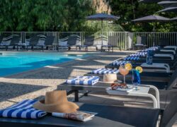 Large pool surrounded by greenery, tiled deck, loungers with towels, colorful cocktails and a book on a low table