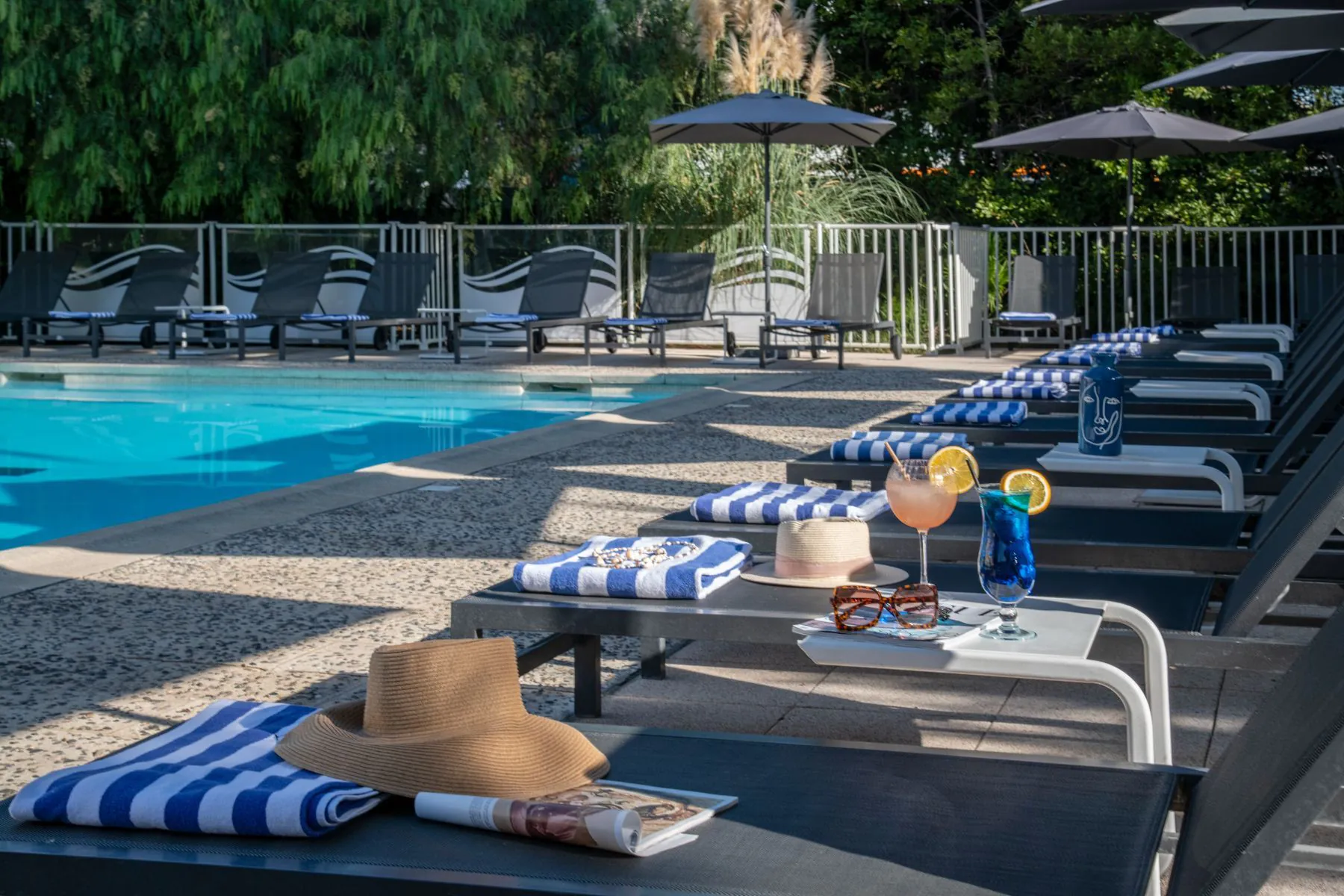 Grande piscine entourée de végétation, terrasse carrelée, transats avec serviettes, cocktails colorés et livre sur table basse
