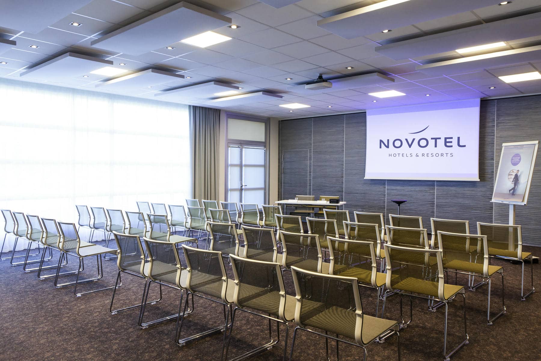 a conference room with rows of chairs and a projector screen that says novotel hotels & resorts