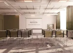 en::Meeting room with sheer curtains and khaki double drapes partially open, thick plum-toned carpet, khaki, black and white chairs arranged in theatre style, facing a screen displaying the Novotel Saint-Quentin-en-Yvelines logo. fr::Salle de réunion avec voilages et doubles rideaux kakis partiellement ouverts, moquette épaisse aux tons parme, chaises kaki, noires et blanches disposées en théâtre, face à un écran affichant le logo Novotel Saint-Quentin-en-Yvelines.