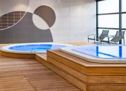 en::Whirlpool area with nearby chairs and coffee tables, wooden flooring, and brown and beige tiled walls featuring soft, rounded geometric patterns. fr::Espace bains bouillonnants avec chaises et tables basses à proximité, sol en bois, murs carrelés marron et beige aux formes géométriques rondes et harmonieuses.