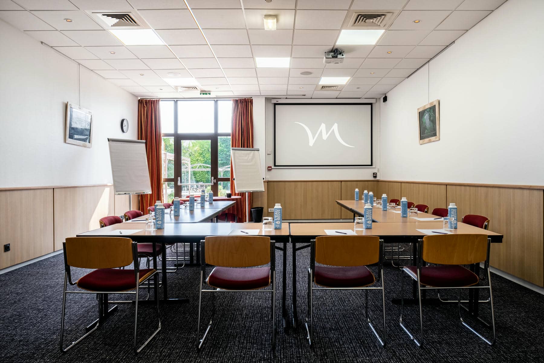 une salle de conférence avec des tables et des chaises et un écran de projection sur lequel figure la lettre m