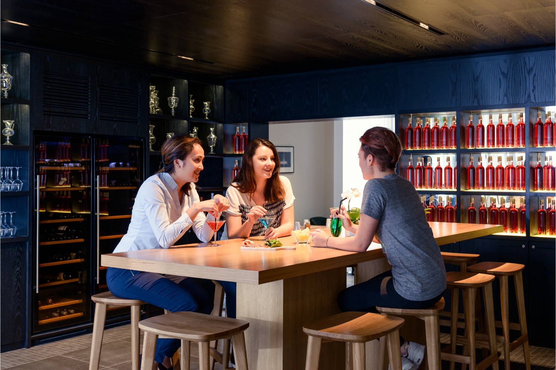 en::Three women chatting over cocktails and a sharing platter at the bar, seated at a wooden high table, with wine cellars and bottle-lined shelves in the background. fr::Trois femmes discutant autour de cocktails et d’une planche à partager au bar, installées à une table haute en bois, avec caves à vin et étagères garnies de bouteilles en arrière-plan.