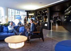 en::Guests seated on the lobby sofas, receptionist welcoming two women in the background, television on showing a sports channel, and corridor leading to the bar and restaurant on the right. fr::Personnes installées dans les canapés du lobby, réceptionniste accueillant deux clientes en arrière-plan, télévision allumée diffusant du sport, et couloir menant au bar-restaurant sur la droite.
