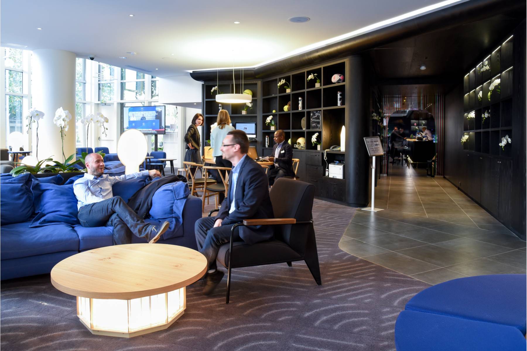 en::Guests seated on the lobby sofas, receptionist welcoming two women in the background, television on showing a sports channel, and corridor leading to the bar and restaurant on the right. fr::Personnes installées dans les canapés du lobby, réceptionniste accueillant deux clientes en arrière-plan, télévision allumée diffusant du sport, et couloir menant au bar-restaurant sur la droite.