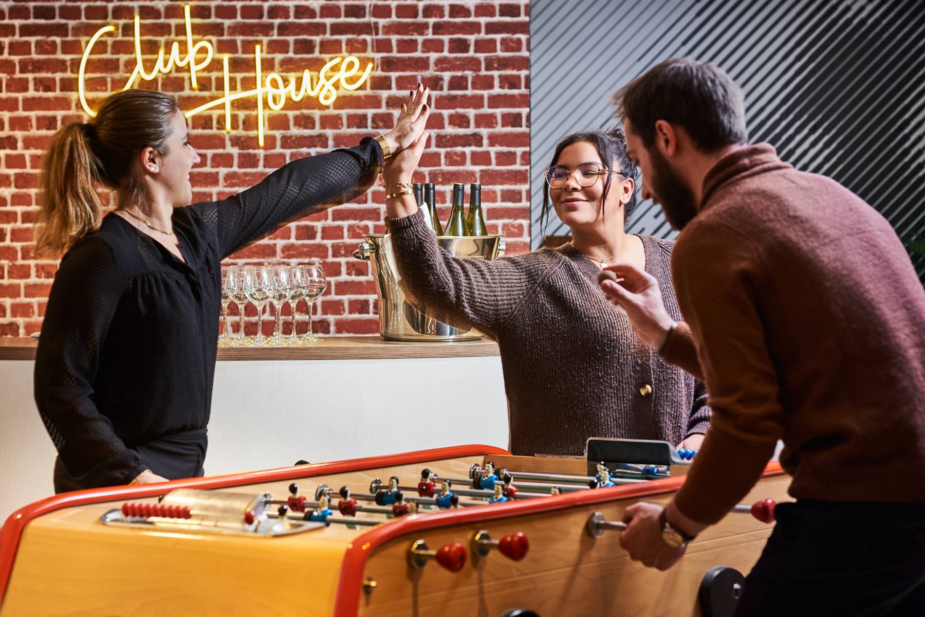 en::Three people playing table football, with a red brick wall and illuminated Club House sign in the background. fr::Trois personnes jouant au babyfoot, avec en arrière-plan un mur de briques rouges et l’enseigne lumineuse Club House