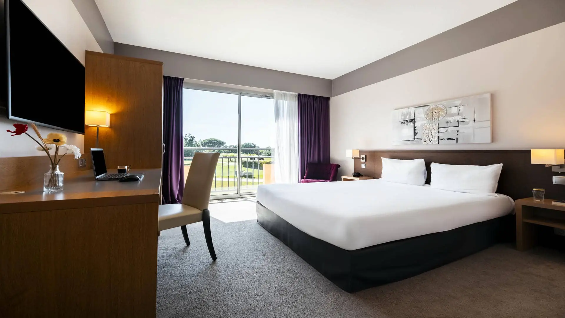 Chambre avec lit double face à un bureau, ordinateur ouvert et café posé à côté, veste noire sur la chaise, avec vue sur la végétation et les bâtiments de l’hôtel séminaire Mercure Toulouse Aéroport Golf de Seilh.
