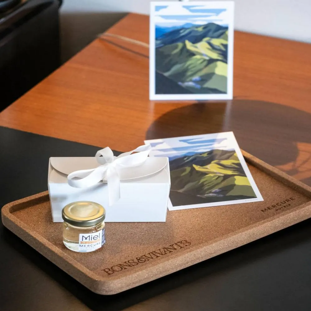A cork tray featuring the Bons & Vivants name, with a jar of honey, a white box tied with a ribbon, and a postcard.
