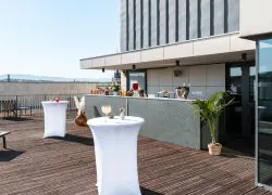 An almond-green metal bar counter set up for a cocktail reception, facing the hotel’s wooden rooftop terrace with views over the city rooftops, complemented by white-covered high tables with floral arrangements, sun loungers, and low tables.