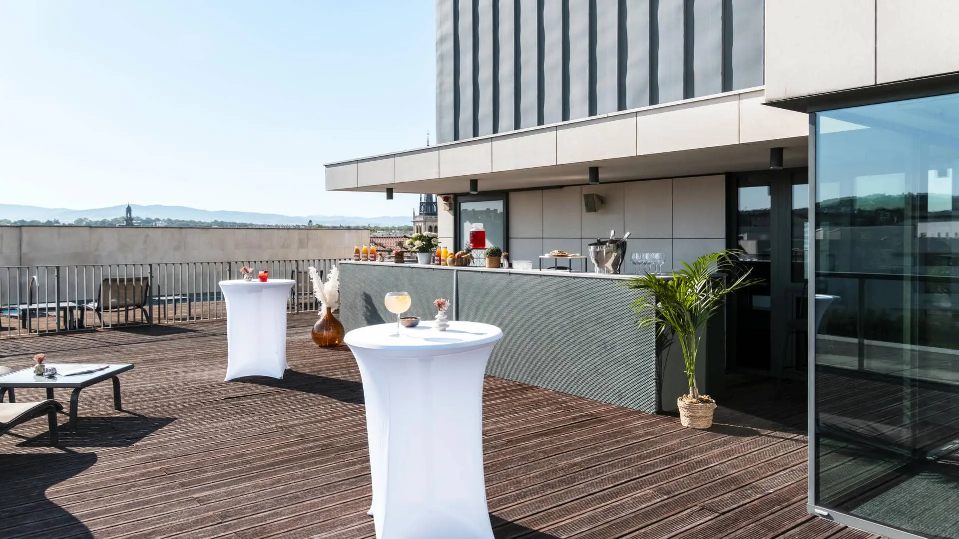 An almond-green metal bar counter set up for a cocktail reception, facing the hotel’s wooden rooftop terrace with views over the city rooftops, complemented by white-covered high tables with floral arrangements, sun loungers, and low tables.