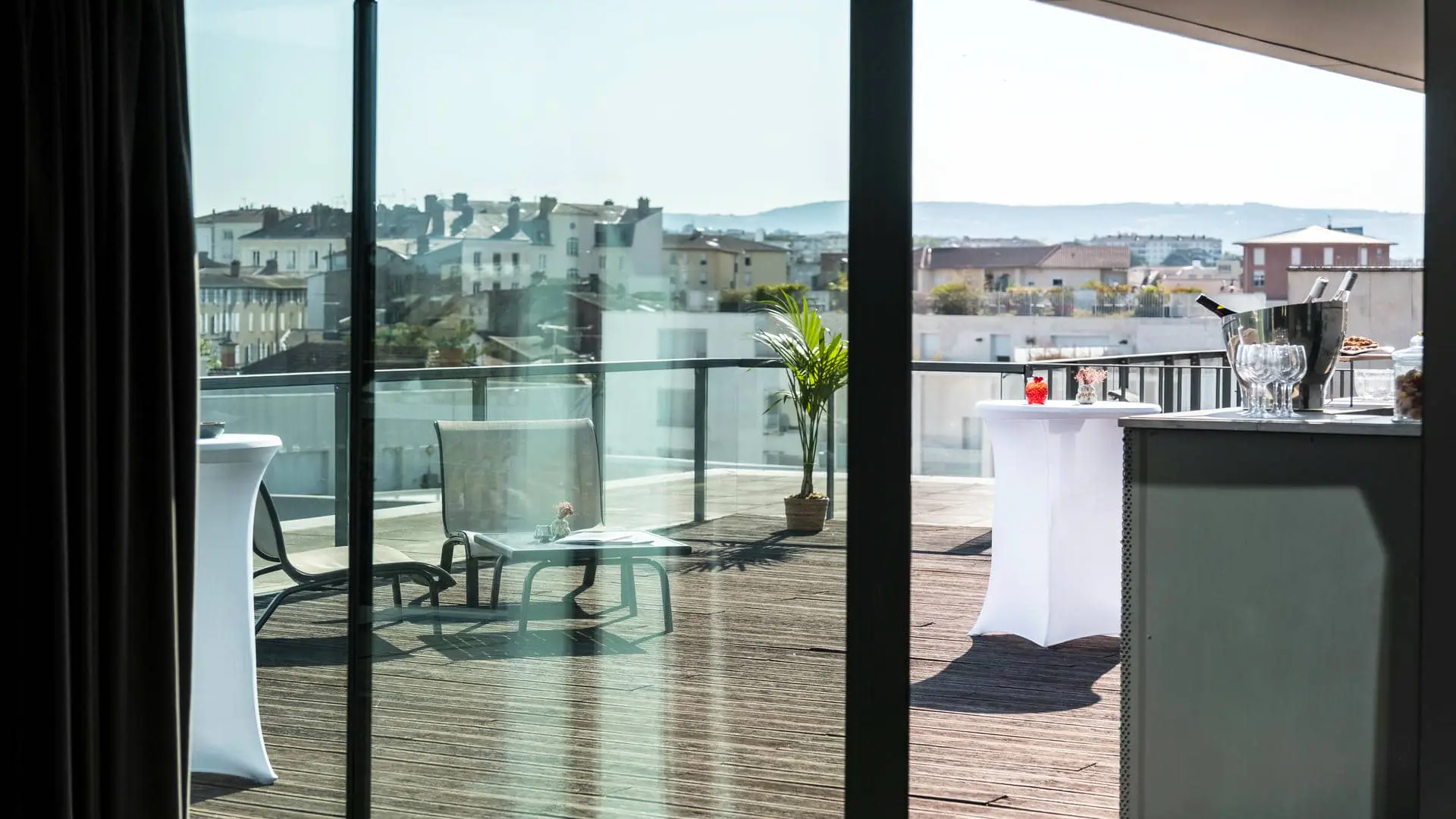 Un comptoir de bar en métal vert amande dressé pour un cocktail, faisant face à la terrasse en bois du rooftop de l'hôtel séminaire avec vue sur les toits de la ville, accompagné de mange-debout habillés de blanc avec compositions florales, de transats et de tables basses.