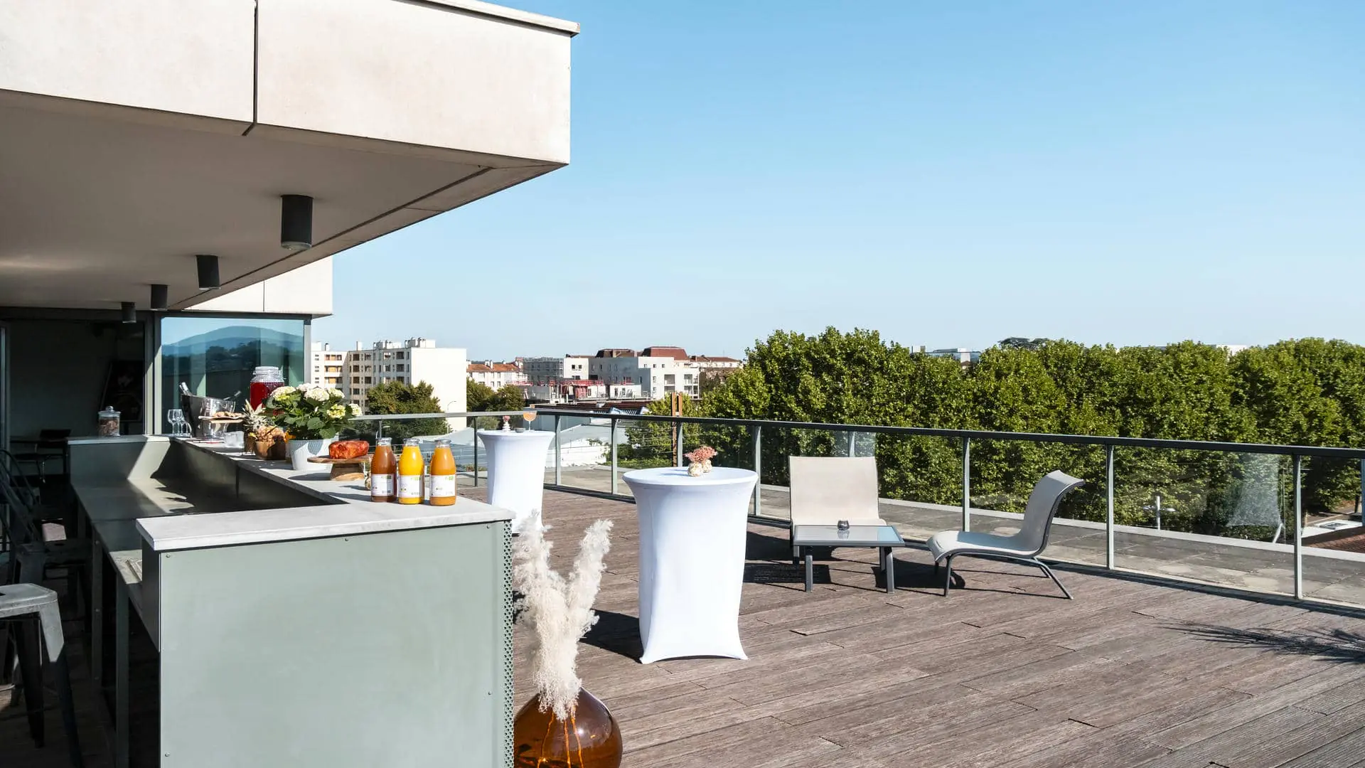 Un comptoir de bar en métal vert amande dressé pour un cocktail, faisant face à la terrasse en bois du rooftop de l'hôtel séminaire avec vue sur les toits de la ville, accompagné de mange-debout habillés de blanc avec compositions florales, de transats et de tables basses.
