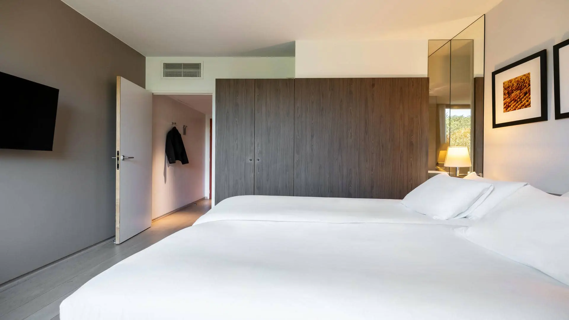 A hotel room with a black wall-mounted television, a brown wardrobe, and a black suit jacket hanging from a wall hook.