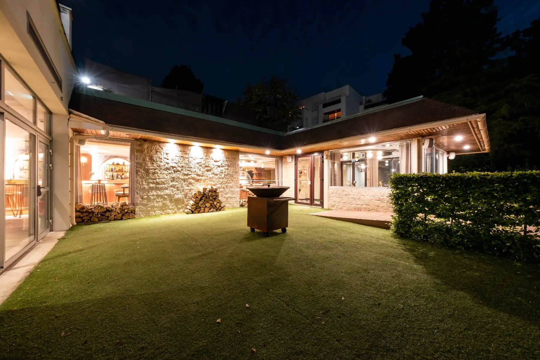 en::Outdoor terrace at night with a fire pit at the center, lawn and shrubs surrounding it, stacked firewood nearby, and the restaurant façade lit in the background. fr::Terrasse extérieure vue de nuit, avec braséro au centre, pelouse et bosquets autour, tas de bois à proximité et façade du restaurant éclairée en arrière-plan.