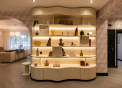 en::White wooden unit with rounded wave-like shapes, featuring backlit shelves displaying vintage travel items such as a trunk, books, and a flask. On the left, a sofa and large screen overlooking the forest; on the right, access to the guest rooms. fr::Meuble en bois blanc aux formes arrondies évoquant des vagues, avec étagères rétroéclairées décorées d’objets de voyage anciens, malle, livres et gourde. À gauche, canapé et grand écran avec vue sur la forêt ; à droite, accès aux chambres.