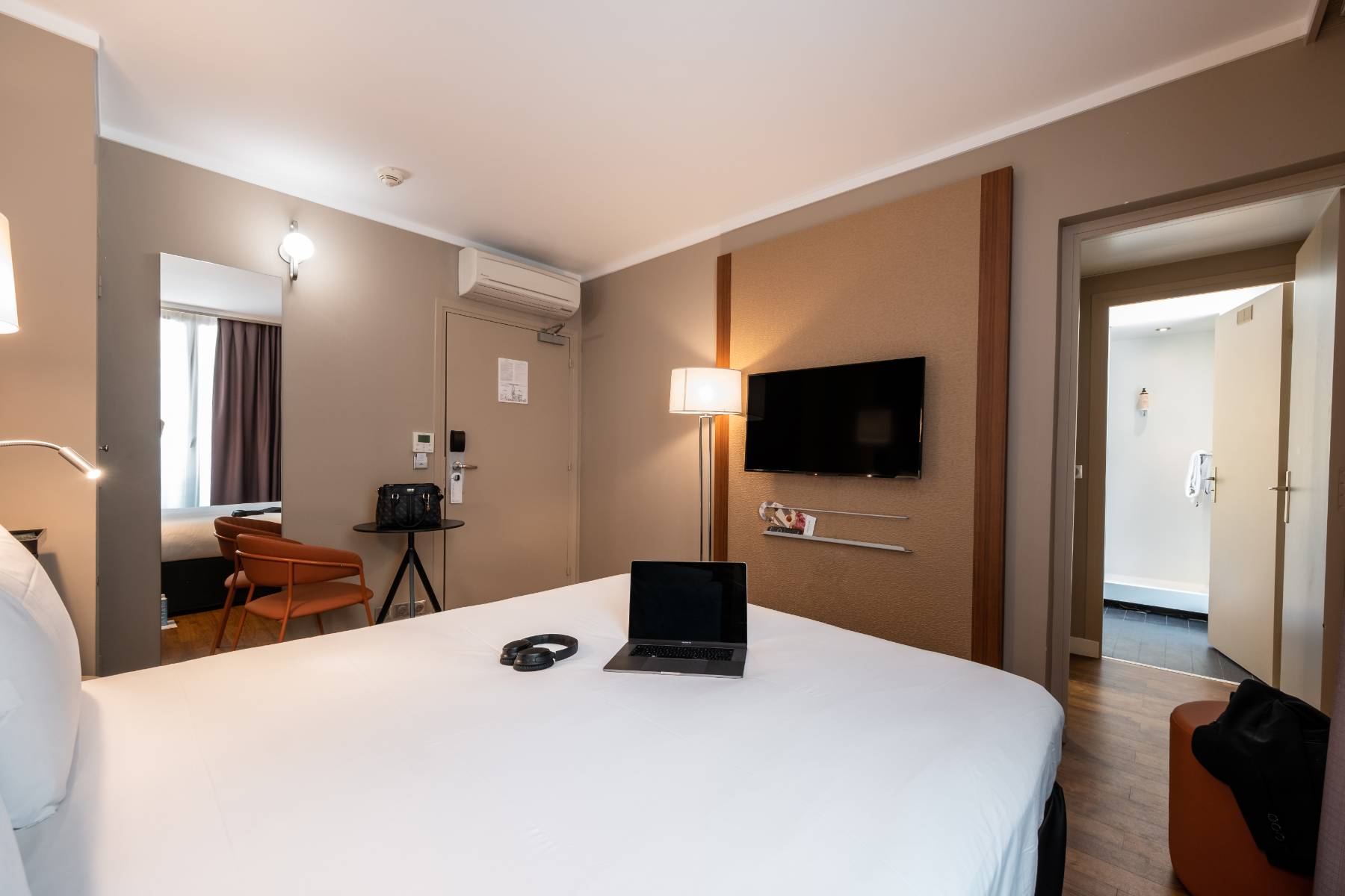 en::Room with a double bed and white duvet, open laptop and headphones placed on it, wall-mounted television, orange fabric chair, and a view of the bathroom. fr::Chambre avec lit double et couette blanche, ordinateur ouvert et casque posés dessus, télévision murale, chaise en tissu orangé et vue sur la salle de bain.
